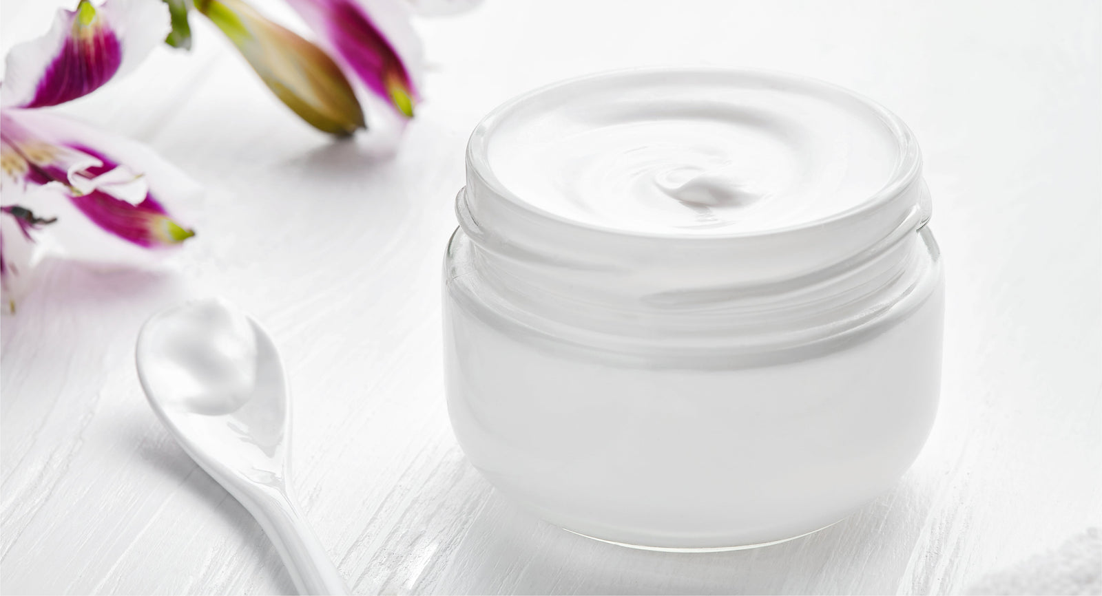An open jar of white cream beside purple flowers.