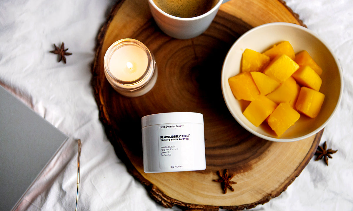 Flawlessly Firm Toning Body Butter on a wooden tray beside mango pieces and coffee.