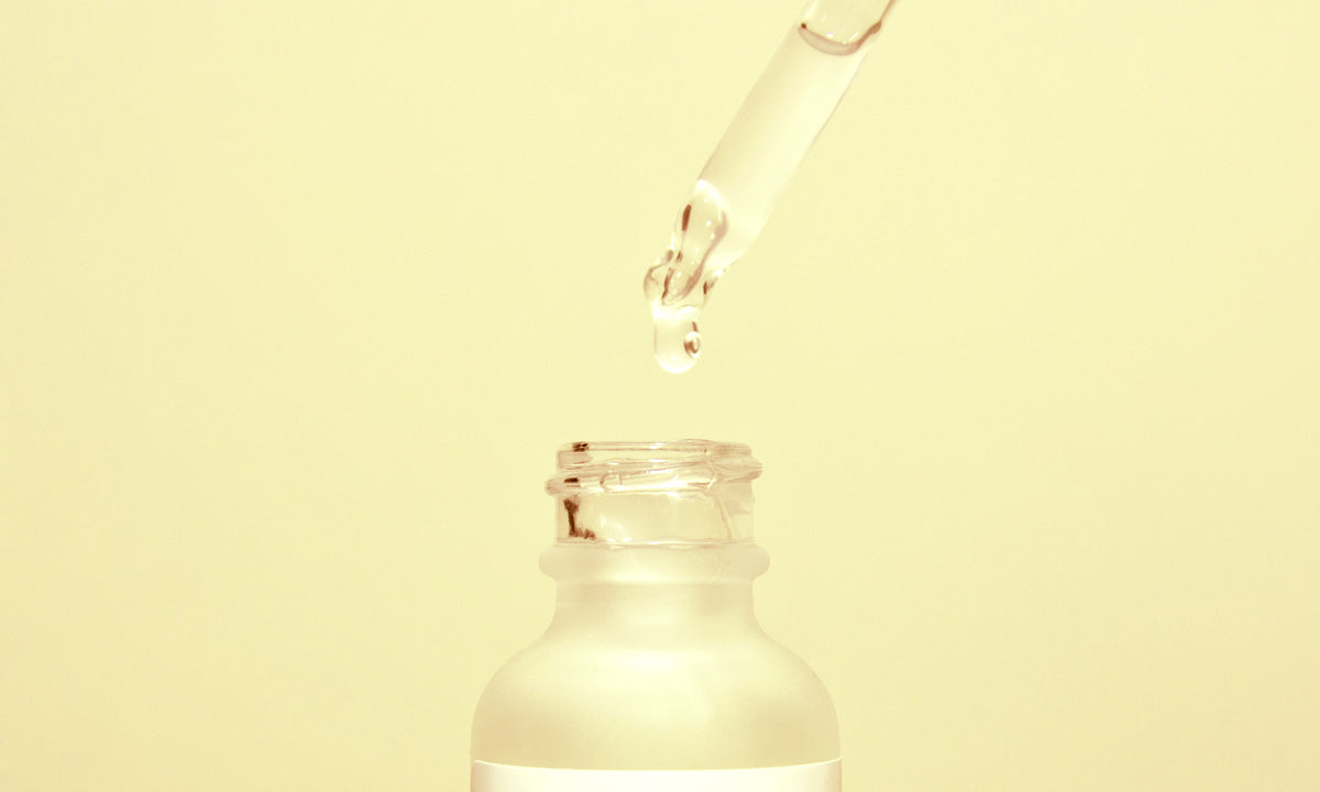 An unbranded frosted serum bottle with a glass dropper above it against a pale gold background.